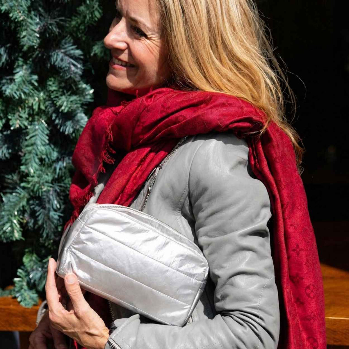 Woman smiling outdoors wearing red scarf and gray jacket, holding silver clutch bag, against greenery background. 