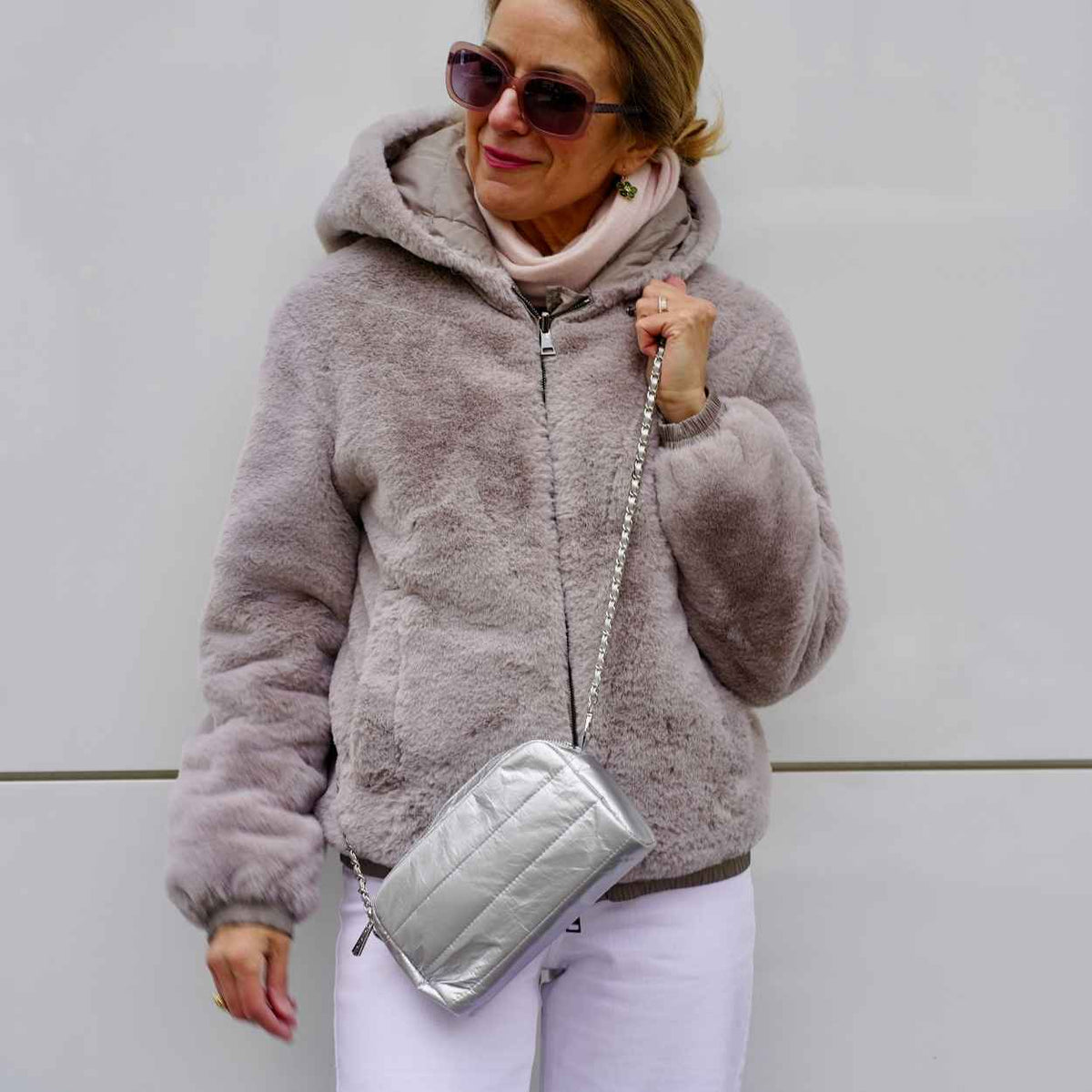 A woman standing against light gray wall in gray fur coat, sunglasses, and wearing a silver mini crossbody purse.