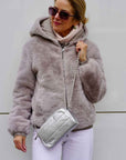 A woman standing against light gray wall in gray fur coat, sunglasses, and wearing a silver mini crossbody purse.