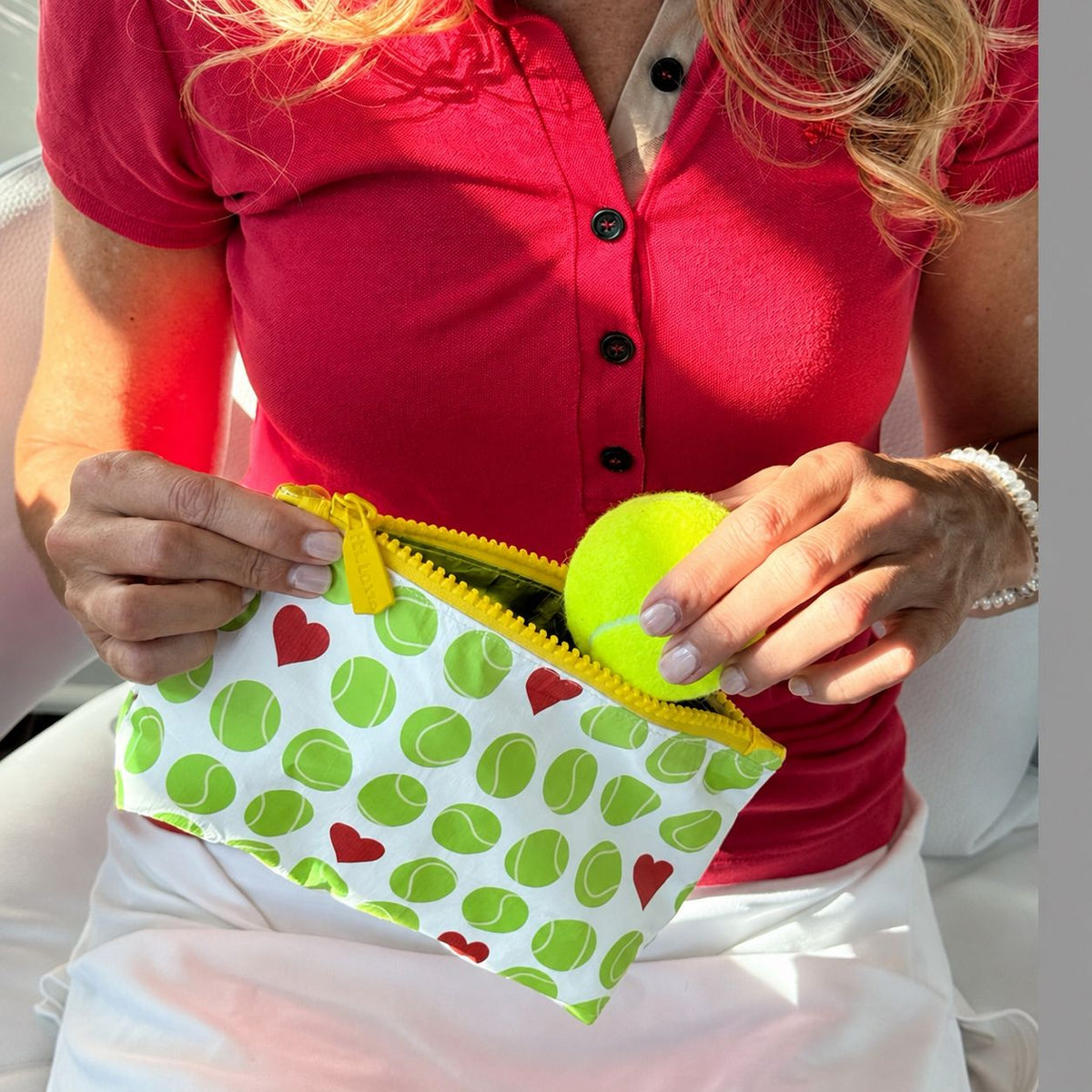 A close-up lifestyle shot of a person holding a white zippered pouch with a green tennis ball pattern and yellow zipper. The pouch is being filled with a bright yellow tennis ball.