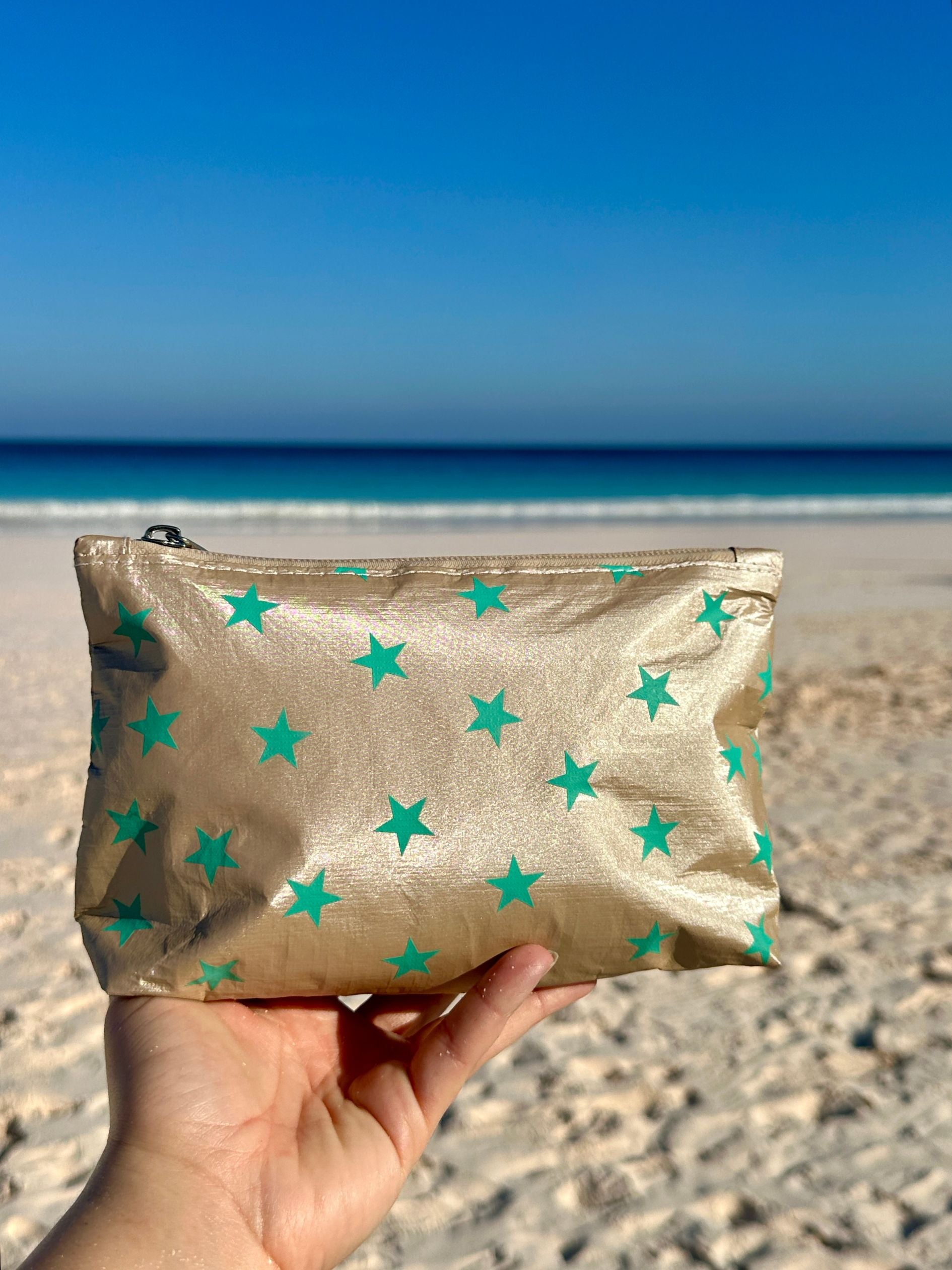 Mini beige zipper pouch with sea green stars in front of ocean