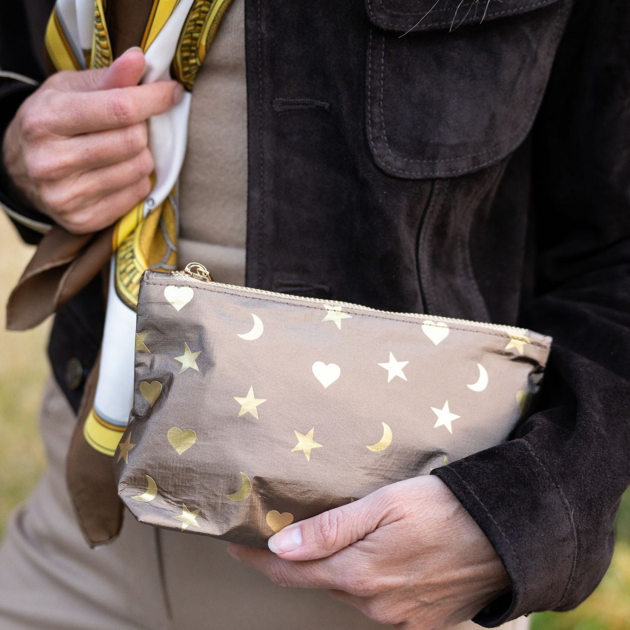 A close-up of a person holding a metallic bronze pouch decorated with gold stars, crescent moons, and hearts. The person is wearing a dark brown suede jacket, a beige shirt, and a yellow and white patterned scarf.