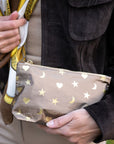 A close-up of a person holding a metallic bronze pouch decorated with gold stars, crescent moons, and hearts. The person is wearing a dark brown suede jacket, a beige shirt, and a yellow and white patterned scarf.