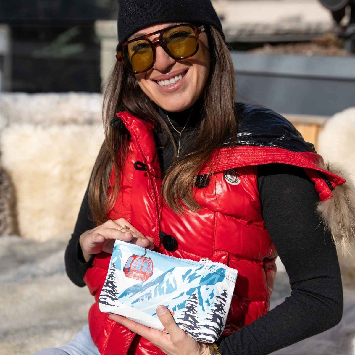 Woman smiling in red vest, sunglasses and hat, holding wristlet with snowy mountain ski design. 