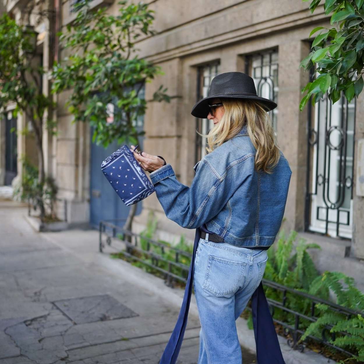 Woman in denim jacket and hat holding a navy bandana pouch on a city street.