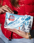 A close up shot of mini zippered pouch featuring a retro-style ski mountain illustration with a red gondola, snowy peaks, evergreen trees, and skis, against red vest. 