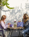 Two women sitting at a table outdoors, each holding a purple bag.