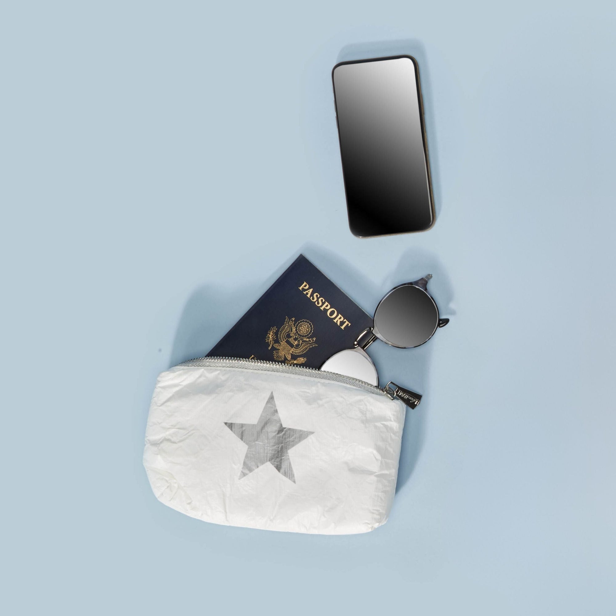 White shimmer mini pouch with silver star holding a phone, sunglasses, and passport, styled as a flat lay on light blue background.