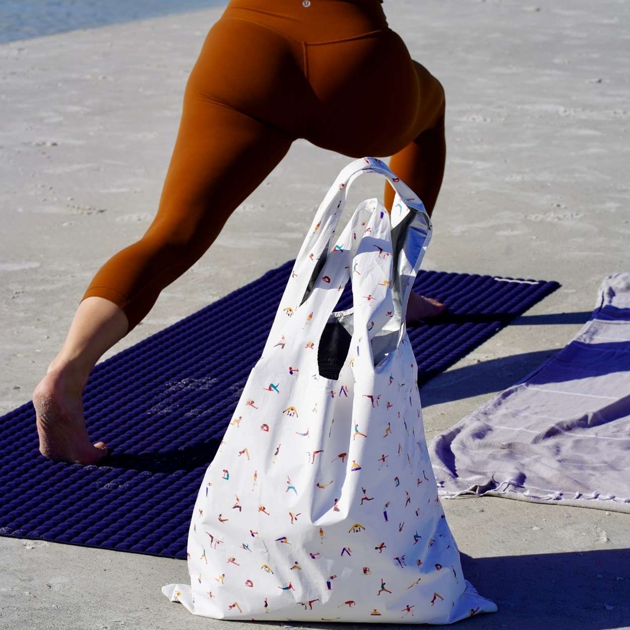 Person on a beach with a white tote bag featuring colorful yoga designs.