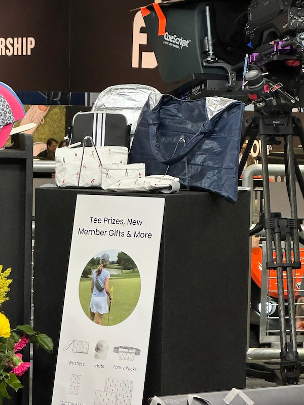 A display table at an event showcasing various Hi Love Travel bags, including a navy blue tote, white patterned bags, and a black and white bag, with a sign below reading 'Tee Prizes, New Member Gifts & More' featured on the Golf Channel.