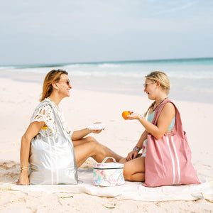A mother and daughter having picnic on the beach. Mom wearing a silver tote bag on her shoulder and daughter on right wearing pink tote bag.