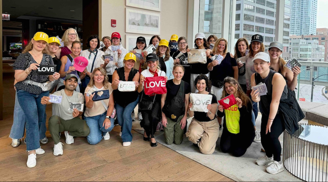 Group of people posing together holding Hi Love Travel Bags in an indoor setting with large windows showing a cityscape.