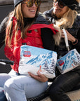Two women holding coordinating zipper pouches with a red gondola and snowy mountain print while aboard a snowmobile.
