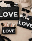 three travel pouches in black with silver LOVE text on beige background with various beauty and travel essentials
