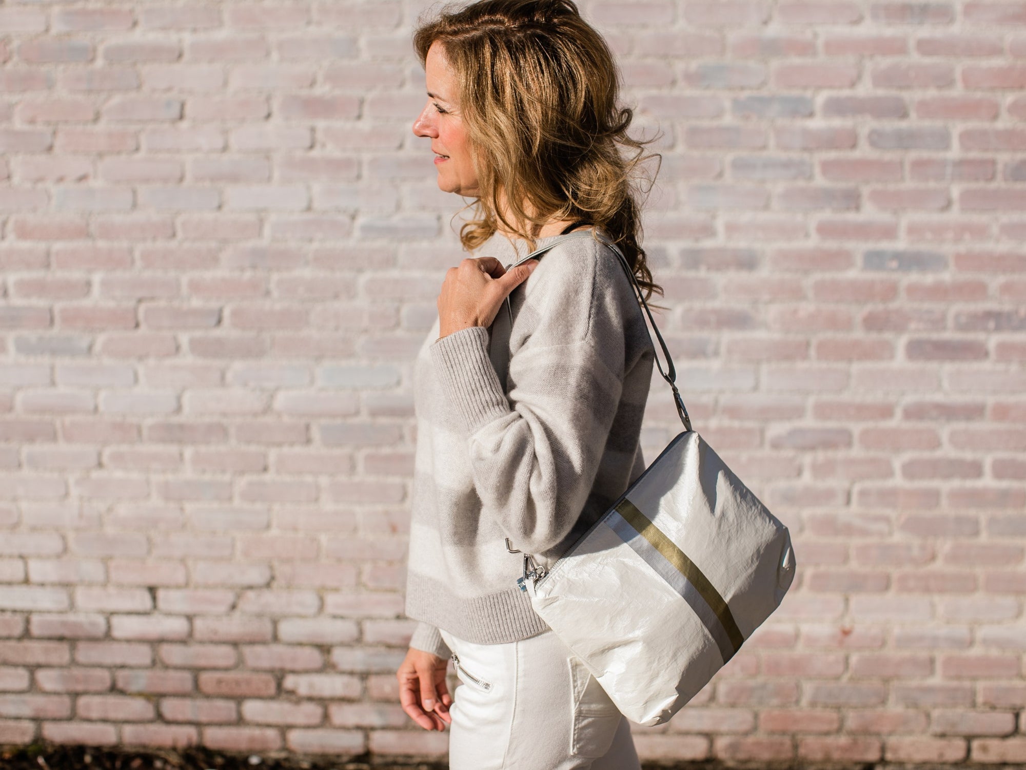 Woman wearing the white shimmer crossbody purse over her shoulder while walking past a brick wall.