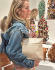 Woman in a denim jacket holding a beige bag in an art gallery with sculptures in the background.