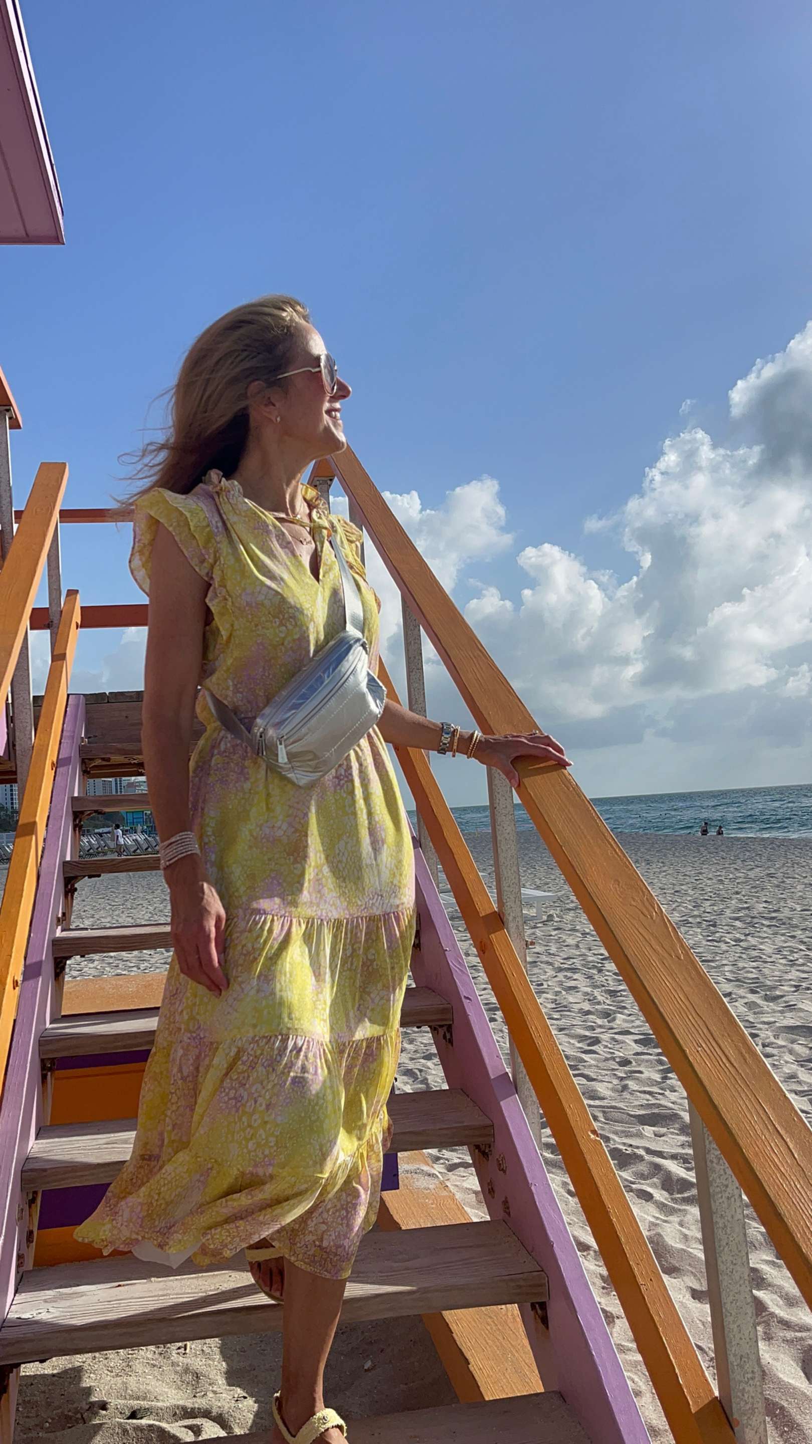 Woman in a yellow dress and silver belt bag standing on a beach with a colorful staircase.
