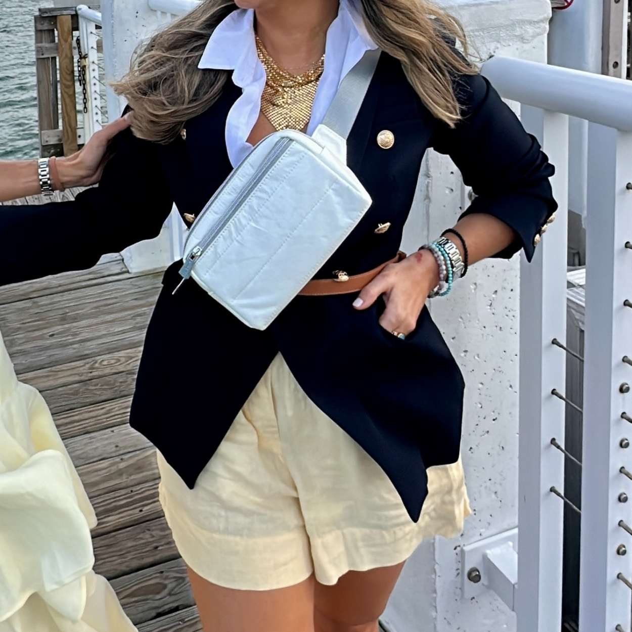 Person wearing a black blazer, white shirt, white fanny pack and beige shorts on a dock.