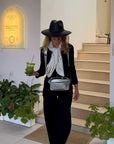 Woman in black outfit with hat, silver fanny pack and scarf, holding a drink, standing in a modern interior setting.