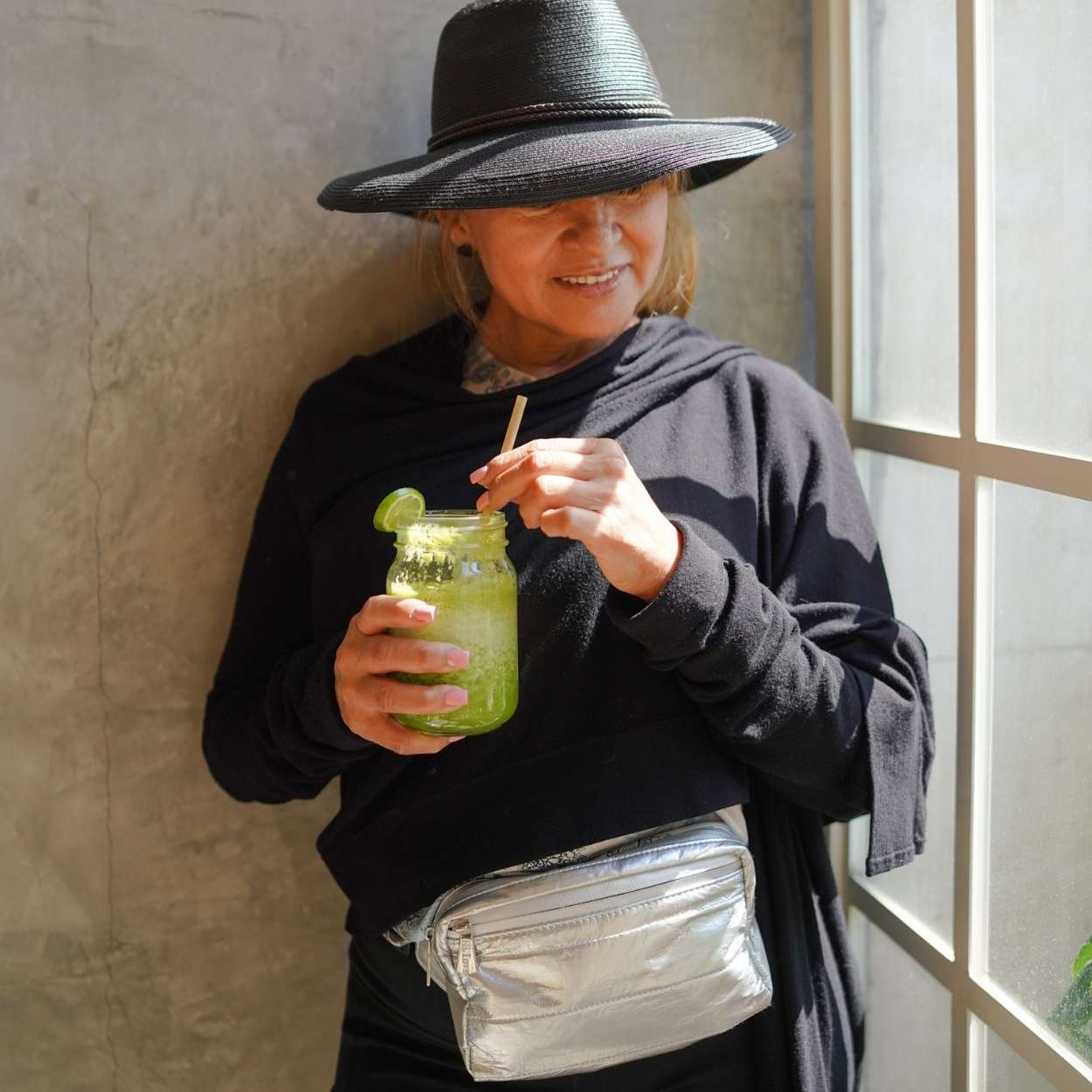 Person holding a green drink with a straw, wearing a black hat, silver fanny pack and black sweater, standing near a window.