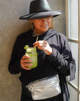 Person holding a green drink with a straw, wearing a black hat, silver fanny pack and black sweater, standing near a window.