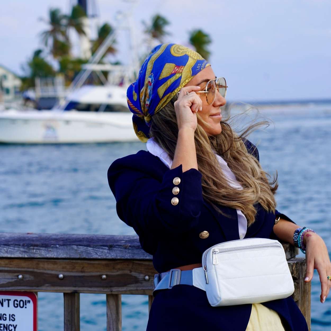 Woman wearing a blue and yellow headscarf, navy jacket, and white bag by a waterfront.