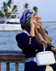 Woman wearing a blue and yellow headscarf, navy jacket, and white bag by a waterfront.