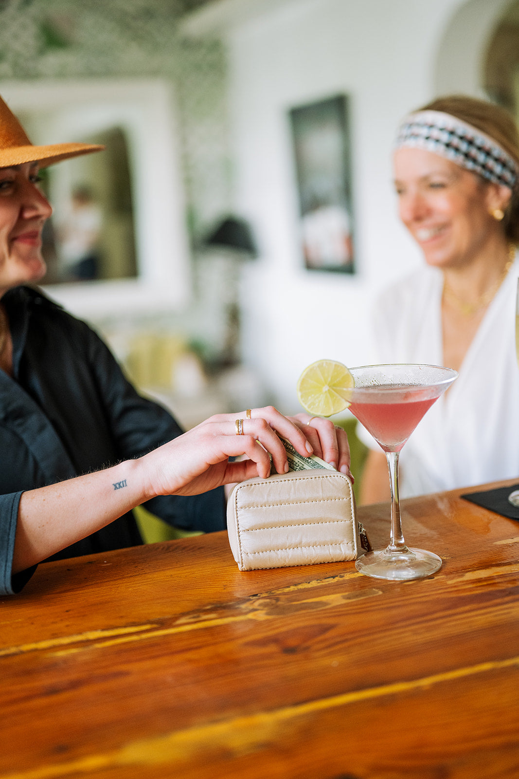 Two smiling women sitting at a bar with beige puffer zipper wallet
