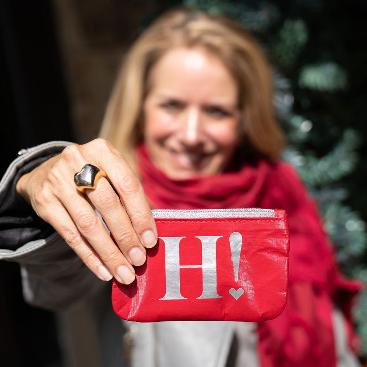 A woman in a red scarf holds up a red card pouch with a large silver letter 'H' and exclamation mark on the front.