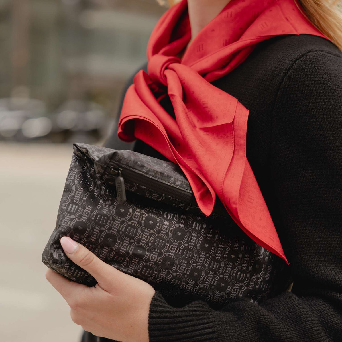 A close-up lifestyle shot of a person holding a black monogram clutch bag with a red H print silk scarf and a black knit sweater.