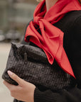 A close-up lifestyle shot of a person holding a black monogram clutch bag with a red H print silk scarf and a black knit sweater.