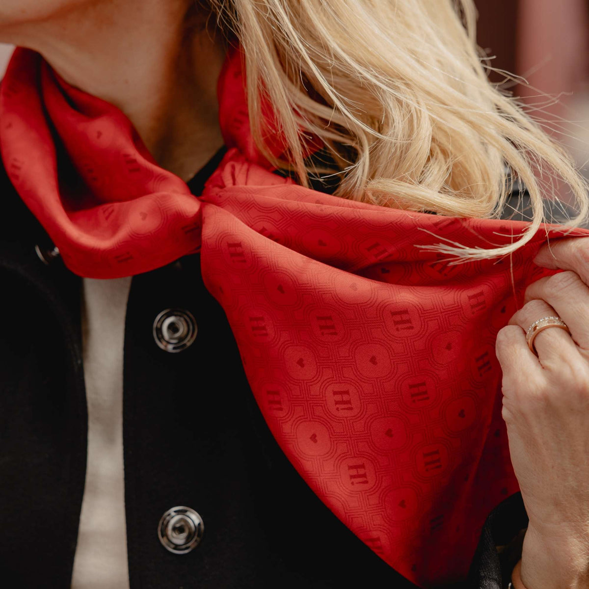 A close-up of a woman wearing a bright red silk scarf with a repeating H signature pattern, styled around her neck over a black jacket.