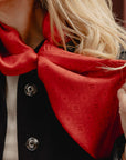 A close-up of a woman wearing a bright red silk scarf with a repeating H signature pattern, styled around her neck over a black jacket.