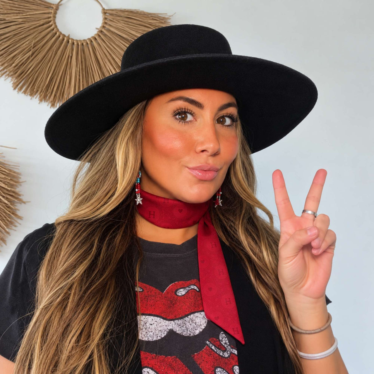 A woman in a black wide-brim hat and graphic T-shirt wearing a deep red tonal scarfette with a signature H-and-heart jacquard pattern, styled snugly around the neck with long pointed ends.