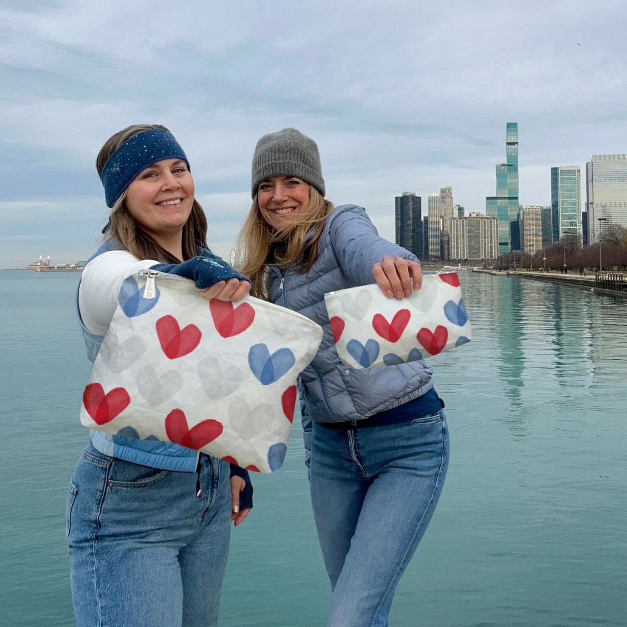 Two women holding bags with heart patterns by a waterfront with a city skyline in the background.