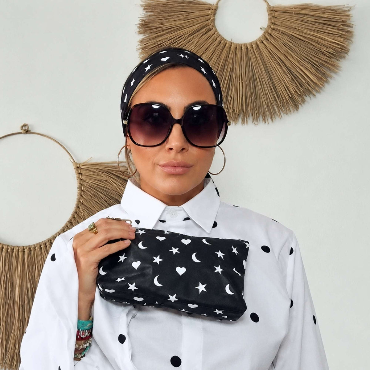 Woman wearing a black silk scarf with hearts, moons and stars as a headband, holding the matching wristlet.