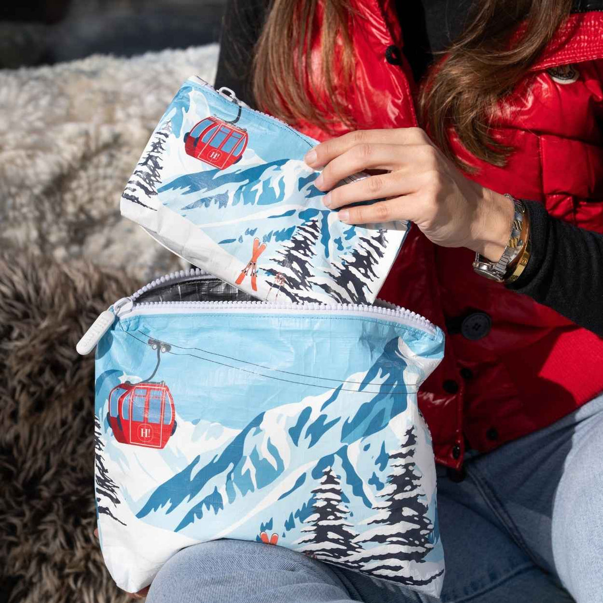 Person in red vest holding two zipper pouches with an alpine snowy mountain scene and red gondola print.