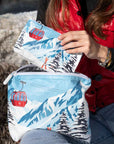 Person in red vest holding two zipper pouches with an alpine snowy mountain scene and red gondola print.