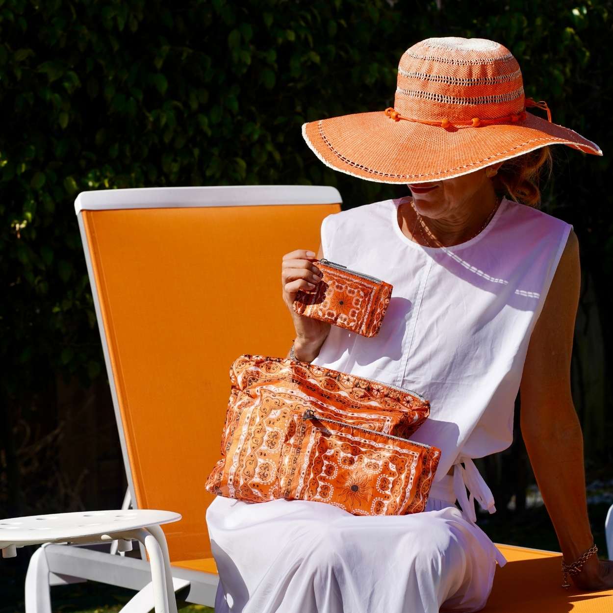 Woman sitting on a chair holding orange bandana patterned bags, wearing an orange sun hat.
