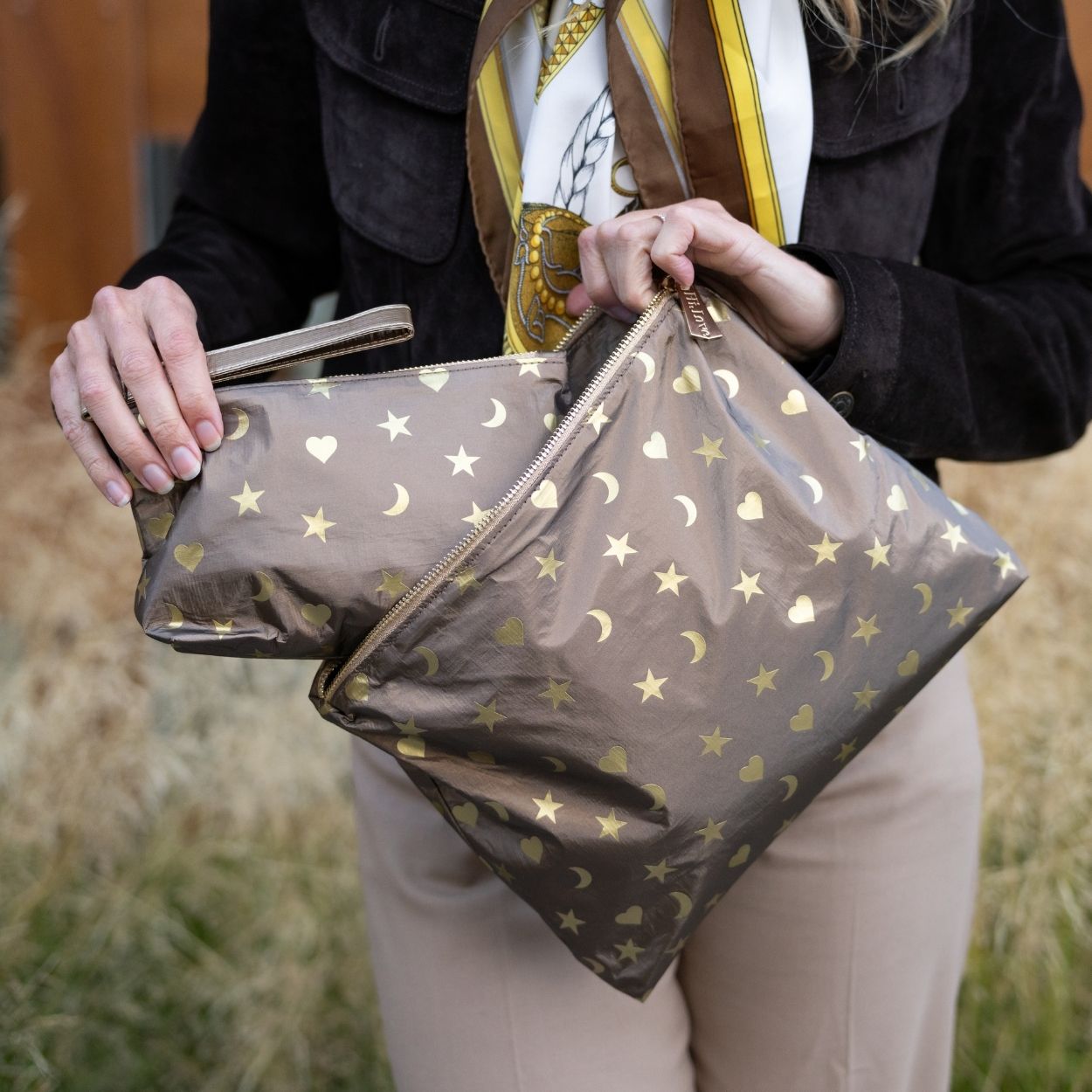 A person outdoors holds metallic bronze zip pouches decorated with gold stars, hearts, and crescent moons. The person is wearing a black jacket, a cream scarf with gold and brown details, and beige pants.
