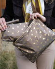A person outdoors holds metallic bronze zip pouches decorated with gold stars, hearts, and crescent moons. The person is wearing a black jacket, a cream scarf with gold and brown details, and beige pants.