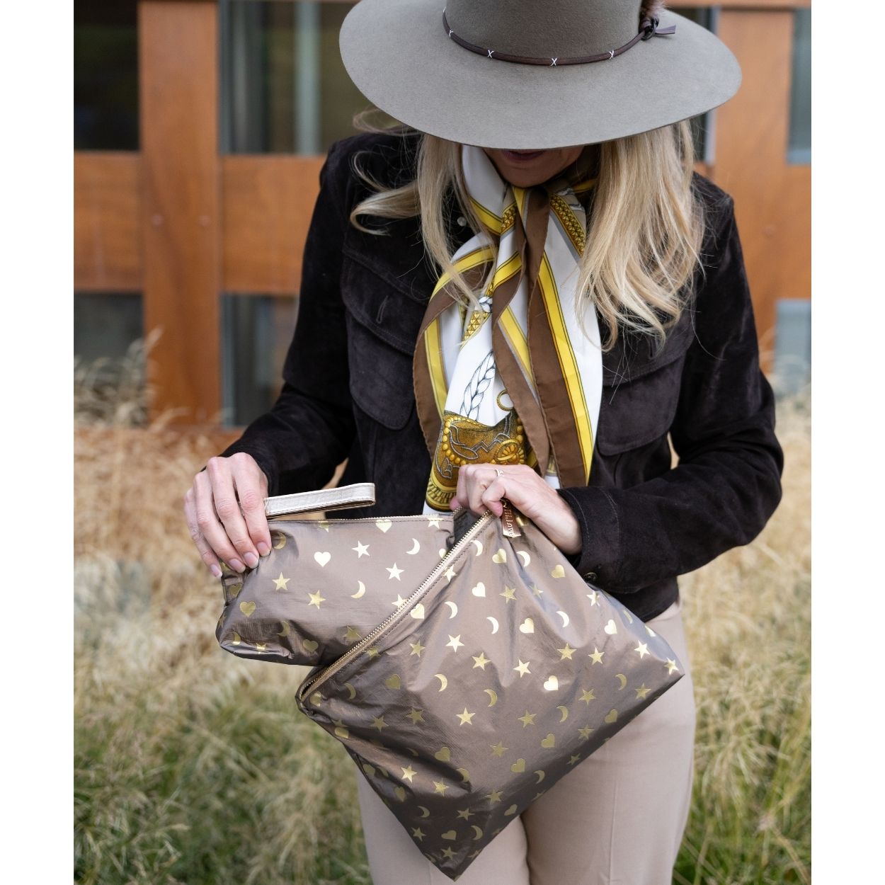 A woman wearing a wide-brimmed taupe hat, dark brown jacket, and a yellow-and-white patterned scarf holds bronze pouches decorated with gold stars, hearts and crescent moons in an outdoor setting.