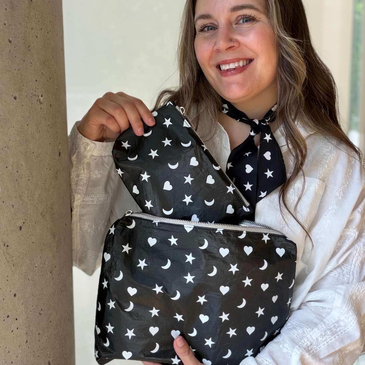 A woman wearing a white lace blouse holds up a black zippered pouch with a white celestial print of stars, moons, and hearts. She is also wearing a matching black neck scarf with the same white pattern.