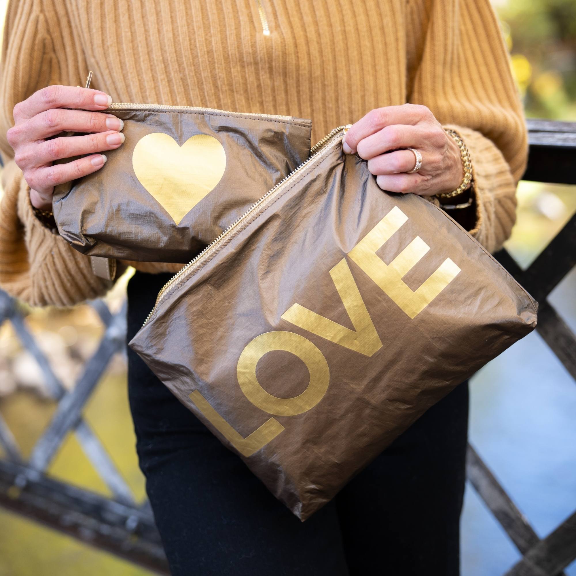 Woman holding a matching set of bronze metallic zipper pouches with oversized gold LOVE lettering and gold heart design in an outdoor autumn setting.