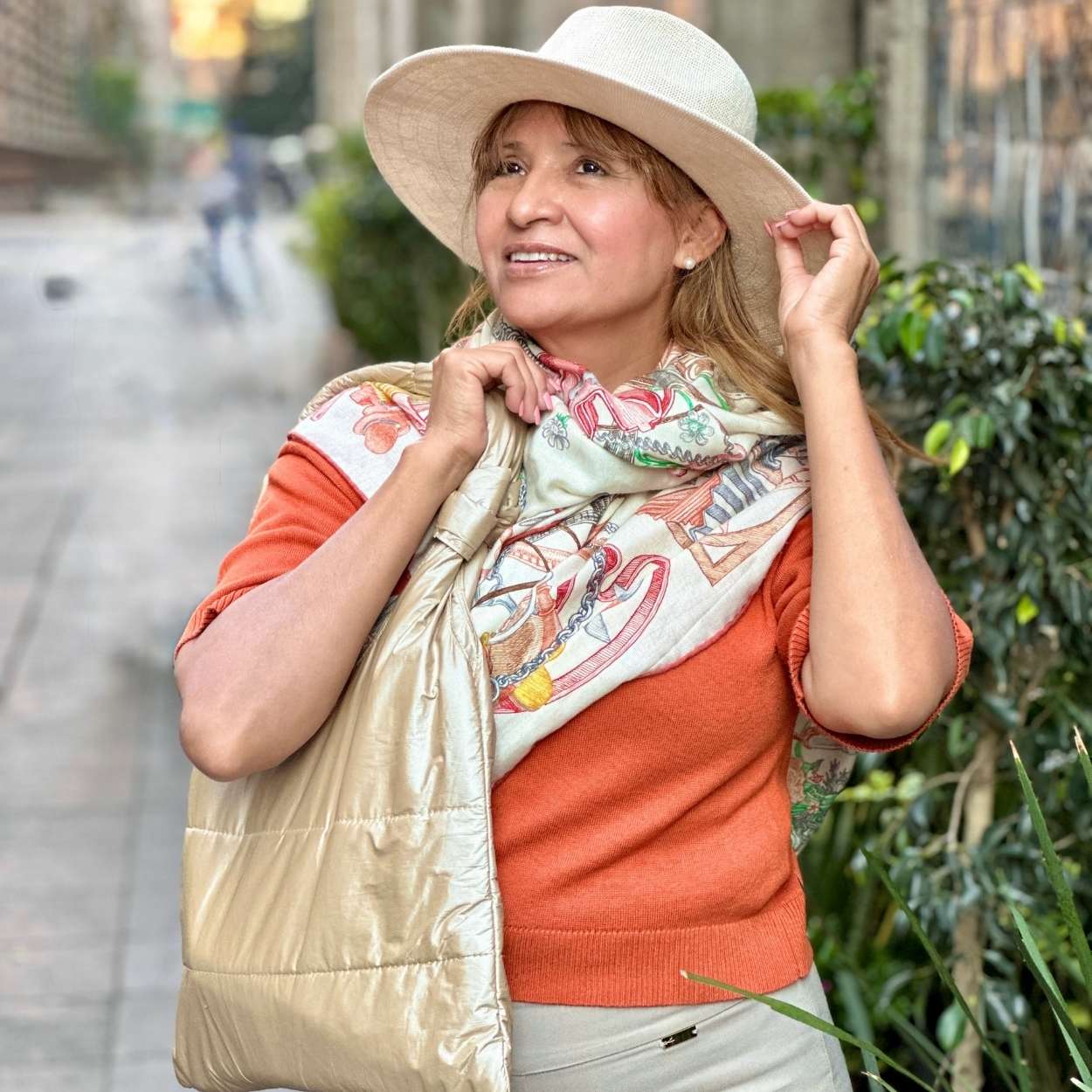 Woman wearing a wide-brimmed hat, orange sweater, and beige puffer tote on a city street.