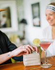 Two smiling women sitting at a bar with beige puffer zipper wallet