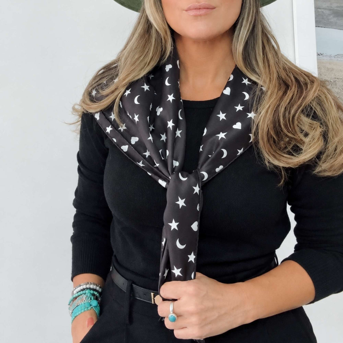 Model wearing a black printed scarf with white hearts, moons and stars tied at the neck, holding the scarf ends.