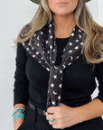 Model wearing a black printed scarf with white hearts, moons and stars tied at the neck, holding the scarf ends.