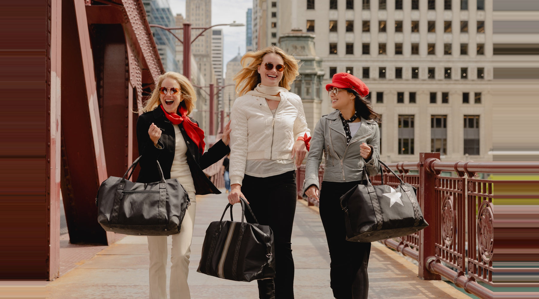 Three women walking hand in hand on a city bridge, each carrying a large black duffel bag. The woman on the left wears a black jacket and white pants with a red scarf, the woman in the center wears a white jacket and black pants, and the woman on the right wears a gray blazer, black pants, and a red beret.