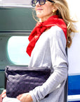 A woman wearing a gray sweater and a red scarf carries a black signature zip pouch in front of an airplane door.